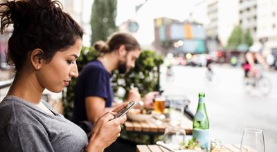 woman sitting at a café looking at her smartphone. Man in the backround is eating. out of focus, bicycles are passing by on the street.